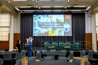 Nicole and Kawal chatting beside the stage before the One King's Impact Summit and Celebration, with the King's College London title slide on the screen behind them and round tables set up for guests