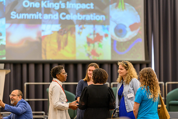 'Funmi speaking to a small group including Katharina, Nicole, and Anica, with student ambassador Chloe in a blue t-shirt nearby and Kawal seated to the left, in front of the One King's Impact Summit a