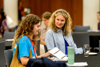 Chloe and Anica seated at a table chatting, with Chloe holding some papers, at the summit event.