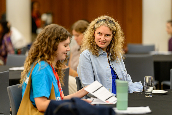 Chloe and Anica seated at a table chatting, with Chloe holding some papers, at the summit event.