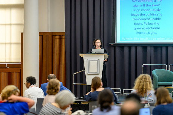 Nicole speaking at the podium to the seated audience during the summit, with a screen behind her displaying instructions.