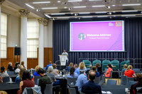 'Funmi delivering the Welcome Address from the podium to a seated audience at round tables, with a slide behind her reading 'Welcome Address, Professor 'Funmi Olonisakin' under the One King's Impact b