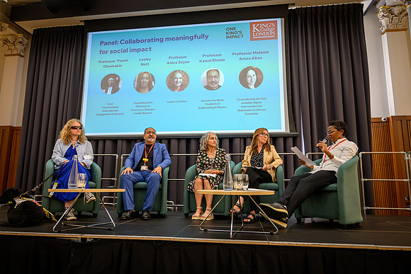 Five panellists seated on stage for 'Collaborating meaningfully for social impact': Anica, Kawal, Melanie, Lesley, and 'Funmi, who is speaking into a microphone as chair, with a guide dog resting besi