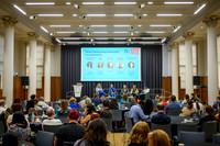 Wide view of the panel discussion 'Collaborating meaningfully for social impact' on stage, with a seated audience at round tables in the foreground and the panellist line-up slide displayed on the lar
