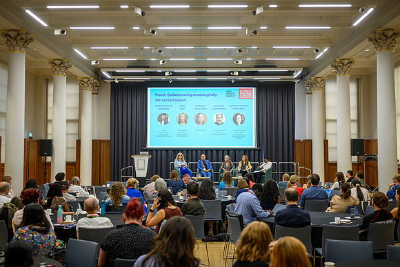 Wide view of the panel discussion 'Collaborating meaningfully for social impact' on stage, with a seated audience at round tables in the foreground and the panellist line-up slide displayed on the lar