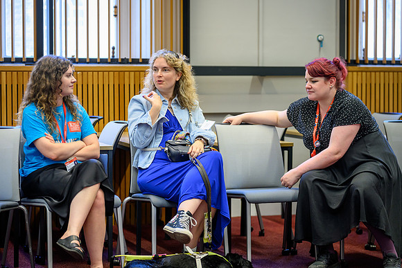 Chloe, Anica, and Anna seated in a small circle chatting during the Community Partnerships Masterclass, with Anica's guide dog resting on the floor beside her.
