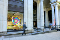 Exterior of Bush House on the Strand, with a colourful circular 'One King's Impact' exhibition graphic in the ground-floor window and a pedestrian passing by, alongside the columned entrance.