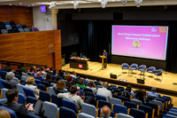 Professor Shitij Kapur, Vice-Chancellor & President of King's College London, delivering the welcome address at a lectern to a full seated auditorium, with empty panel chairs set up on stage and a scr